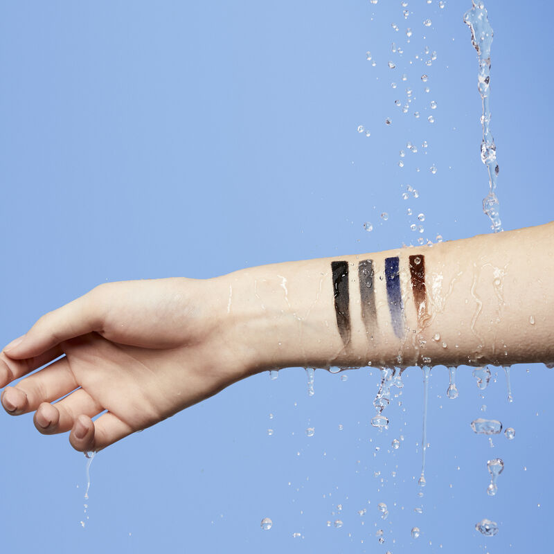 Fair skinned arm with gradient bars of Mary Kay Waterproof Eyeliner in Black, Steely, Blue and Brown being drenched in water against blue background.